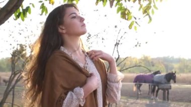 Uzun saçlı model kadın gün batımında atların yanında duruyor.