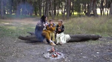 Happy family mother and two young kids sitting on tree in park near camp fire in autumn