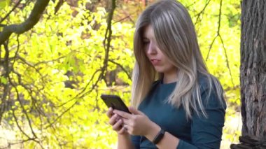 Sonbahar parkında sonbahar yaprakları arasında akıllı telefon kullanan mutlu kadın. 