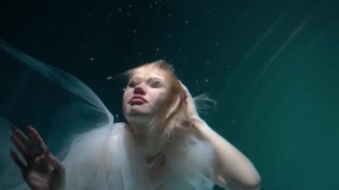 Beautiful blonde girl in a white dress swims underwater in a pool with bubbles