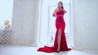 Young adult elegant woman in red long dress standing near window 