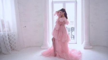 Pembe elbiseli hamile kadın beyaz odada el ele tutuşup poz veriyor.