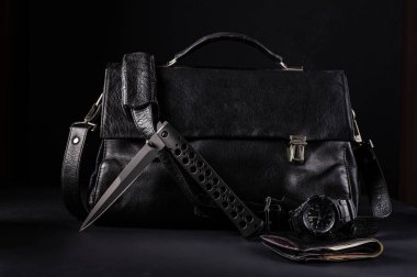 Siyah deri bir çantanın arka planında büyük siyah bir katlanır bıçak. Beyefendiler bıçak, saat ve cüzdan seti. Bıçak yarı katlanmış pozisyonda..