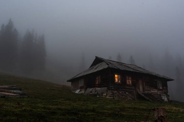 Lonely hut in the forest. Lonely wooden house in the mountains. Gloomy house with luminous windows. Fog in the mountains. clipart
