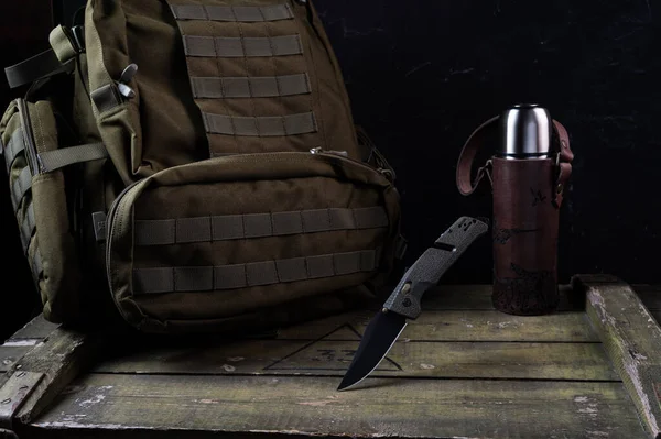 Army knife with black blade and olive handle. Green knife for the military and a green backpack with a molle system. Thermos in a leather case.