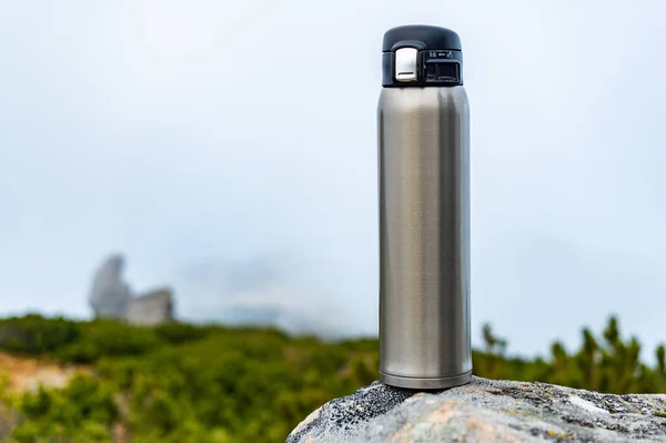 Steel thermos on a stone. Steel bottle on a stone in the mountains. Bottle on the background of clouds and mountains. Front view.