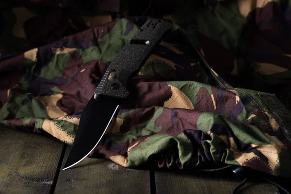 Army knife with black blade. Knife and army uniform. Camouflage and green box. Front view.