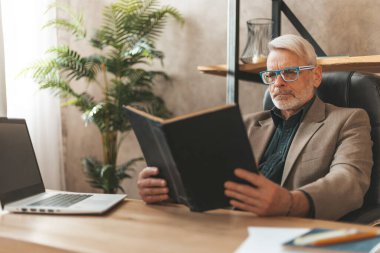 Gözlüklü ve sakallı yaşlı bir adam masada bir kitap tutuyor. Boş vakitlerinde sakin bir şekilde okuyabilirsin. Yaşlılıkta öğrenme ve yeni bilgi.