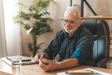 A mature man measures his blood pressure, hypertension in the elderly. Cardiac check at home with a stethoscope.