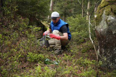Yetişkin adam kırmızı eldivenli gönüllü ormandaki plastik çöpleri temizliyor, çevreyle ilgileniyor..