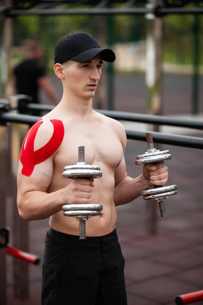 Young European athlete with injury is training outdoors. Kinesiology ( therapeutic, Kinesio, k-tape, KT, physio) tape on shoulder. Rehabilitation and recovery. Exercise with dumbbells.