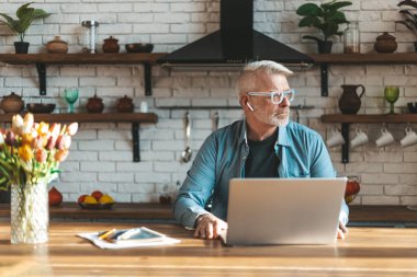 Ofis mutfakta. Kıdemli adam elinde dizüstü bilgisayarla bir masada oturuyor. Evde uzaktan çalışma.