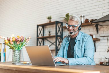 Kıdemli blogcu. Gri saçlı bir adam dizüstü bilgisayarlı bir masada gülümsüyor. Video çağrısı. İç mimari.