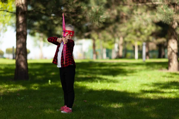 Archer child. Little girl takes aim with a bow in hands in the park outdoors. Superhero game.