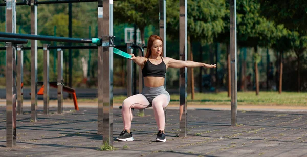 Strong woman squats with an elastic band. Exercises on urban sports ground.