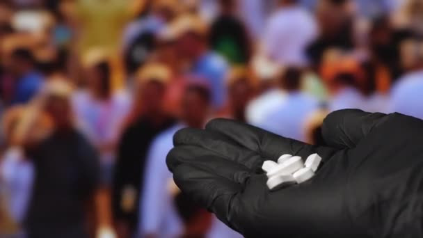 Una mano en guantes de látex negro sosteniendo píldoras blancas con personas en el fondo. — Vídeos de Stock