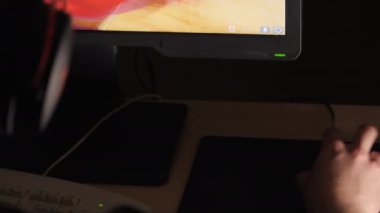 Karanlık bir odada bilgisayar oyunu oynayan bir adamın yakın plan fotoğrafı. Bej masa. Beyaz ırk kökenli. Modern teknoloji. Klavye ve fare kullanarak. Kulaklık takıyordu. Göster. Seçici odak.