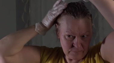 Caucasian lady coloring her hair. Using hair dye. Red hair. Middle-aged. White and gray background. Yellow shirt. White rubber gloves. Camera slowly moving around.