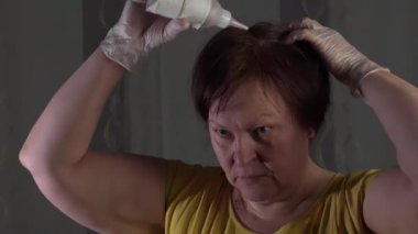 Caucasian lady styling her hair. White rubber gloves. Yellow shirt. White and gray background. Middle-aged. Red hair. Using hair dye. Camera slowly moving around.