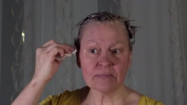 Orta yaşlı beyaz kadın saçını boyattıktan sonra yüzünü temizliyor. Beyaz ve gri arka plan. Kızıl saçlı. Sarı gömlek. Kamera yavaşça hareket ediyor.
