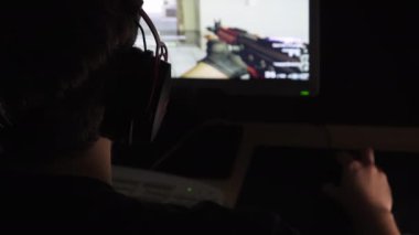 Çocuk karanlık bir odada bilgisayar oyunları oynuyor. Modern teknoloji. Bej masa. Beyaz ırk kökenli. Göster. Klavye ve fare kullanarak. Kulaklık takıyor..