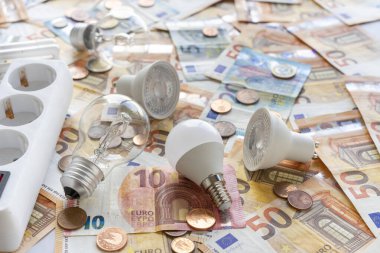 Elektrik maliyetleri: Avro banknotları ve sikkelerin arka planında çeşitli ampul türleri ve elektrik soketi. 