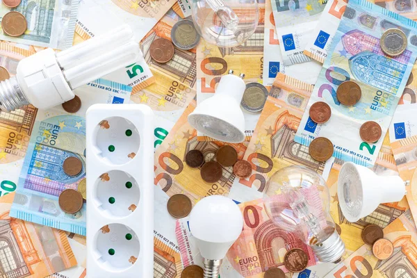 Elektrik maliyetleri: Avro banknotları ve sikkelerin arka planında çeşitli ampul türleri ve elektrik soketi. 