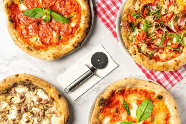 Çeşitli pizza türlerinin üst görüntüsü: Prosciutto 'lu pizza, mantarlı pizza, salamlı pizza, kesme aracıyla kesilmeye hazır, beyaz arka plan ve kırmızı kareli masa örtüsü..