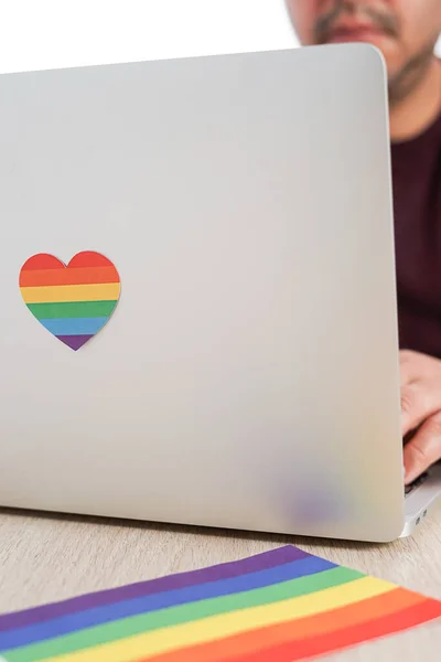 Uomo ispanico che lavora con il suo computer che ha un cuore con i colori LGBTQ e sul tavolo la bandiera del movimento. Concetto di tolleranza e diversità. — Foto Stock