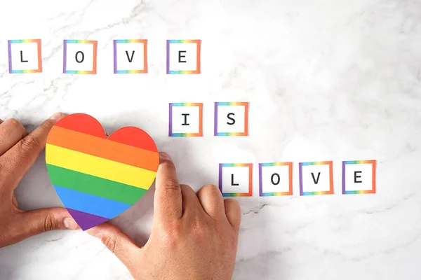 Hands of a man placing a Heart with the LGBTQ colors with the phrase Love is Love on a marble background. Concept tolerance, diversity and inclusion. — Stockfoto