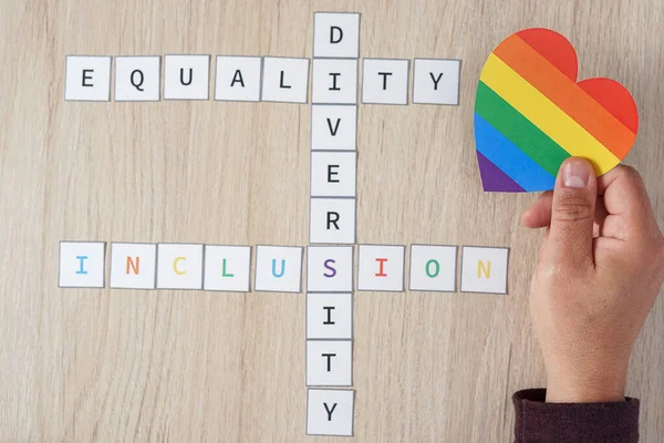 Um homem maduro mão colocando um coração com cores LGBTQ ao lado das palavras igualdade, diversidade e inclusão em um fundo de madeira. Conceito de tolerância e diversidade. — Fotografia de Stock