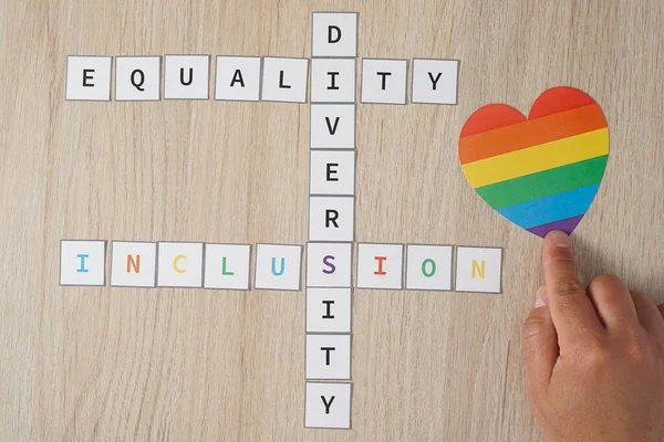 Uma mão de homem colocando um coração com cores LGBT ao lado das palavras igualdade, diversidade e inclusão em um fundo de madeira. Conceito de tolerância e diversidade. — Fotografia de Stock