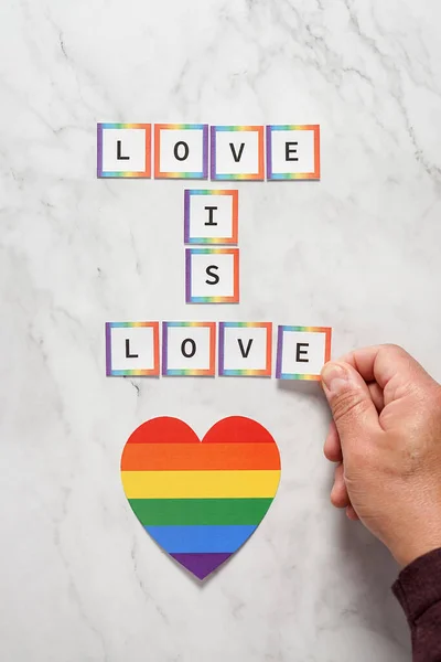Uma mão de homem terminando a última letra da frase Amor é Amor ao lado de um coração com as cores do movimento LGBTQ. Conceito de tolerância, inclusão e diversidade — Fotografia de Stock