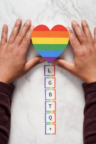 Mãos de um homem maduro colocando um coração com as cores do movimento sobre o acrônimo LGBTQ. Conceito de tolerância, inclusão e diversidade — Fotografia de Stock