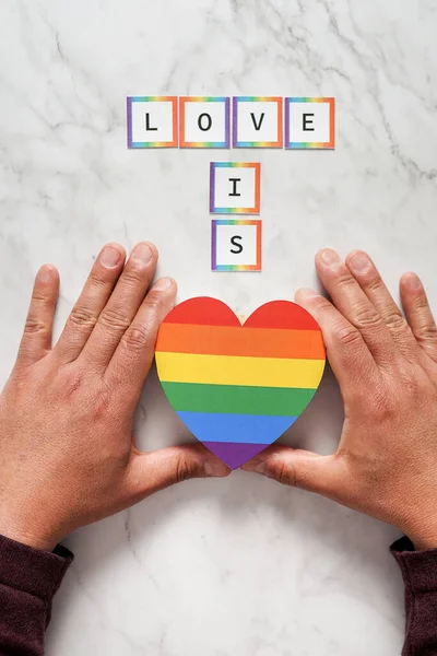 Hands of a mature man placing a heart with the colors of the LGBTQ movement under the phrase Love is. Concept of tolerance, inclusion and diversity — Stockfoto