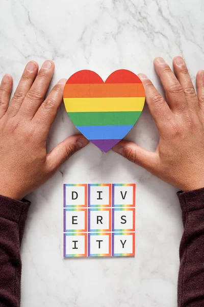 Ruce dospělého muže umísťujícího srdce s barvami hnutí LGBTQ nad slovem rozmanitost. Koncept tolerance, inkluze a rozmanitosti — Stock fotografie
