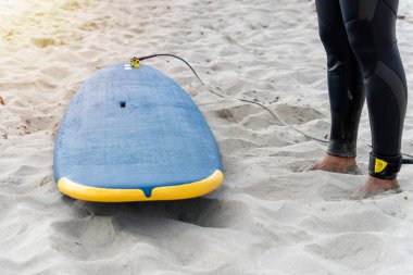 Surfboard on the beach sand next to the legs of an unrecognizable surfer on a sunny day. High quality photo