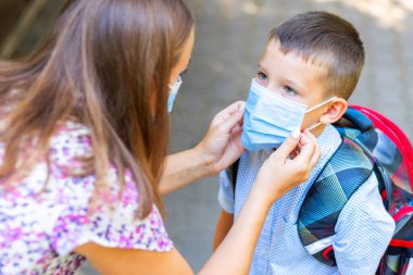Tıbbi maskeli küçük bir çocuk ve anne. Anne oğullarının yüzüne koruyucu bir maske takıyor. Öğrenci çocuk okula gitmeye hazır. Dışarıda sırt çantası olan sevimli bir çocuk. Okul konseptine dönelim..