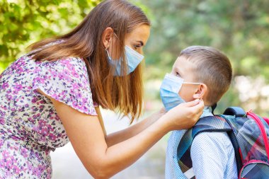 Tıbbi maskeli küçük bir çocuk ve anne. Anne oğullarının yüzüne koruyucu bir maske takıyor. Öğrenci çocuk okula gitmeye hazır. Dışarıda sırt çantası olan sevimli bir çocuk. Okul konseptine dönelim..