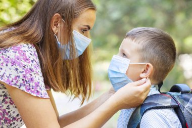 Tıbbi maskeli küçük bir çocuk ve anne. Anne oğullarının yüzüne koruyucu bir maske takıyor. Öğrenci çocuk okula gitmeye hazır. Dışarıda sırt çantası olan sevimli bir çocuk. Okul konseptine dönelim..