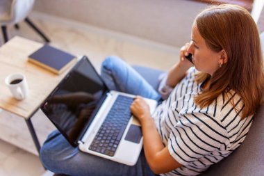 Evdeki kanepede dizüstü bilgisayarla otururken cep telefonu kullanan mutlu genç bir kadın. Bir kadın cep telefonuyla konuşuyor, sorular soruyor, bilgisayar ekranına bakıyor..