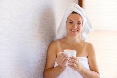 Banyo havlusu içinde çekici bir genç kız ve kafasında bir havluyla bir fincan tutuyor ve gülümsüyor..