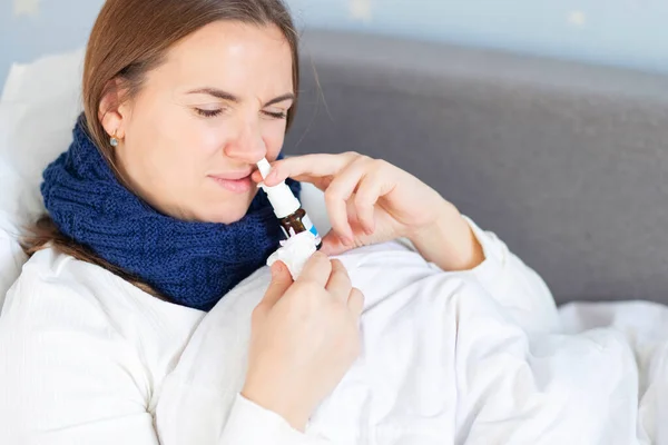 A young woman with bad cold lying in bed under the blanket and using nasal drops. Woman using nose spray.