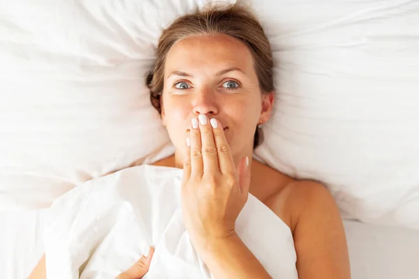 Young woman lies in bed and covers her mouth with her hand. Womens secrets concept.