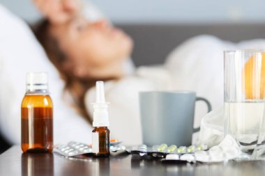 Yatağında yüksek ateş ve grip yüzünden battaniyeyle yatan hasta genç bir kadın. Yatak odasında bir sürü ilaç var..