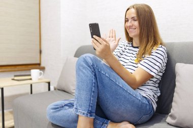 Cheerful young woman is using smartphone for video call, gesturing hi to friend or parent. Smiling young caucasian woman sits relax on couch in living room talks on video call.