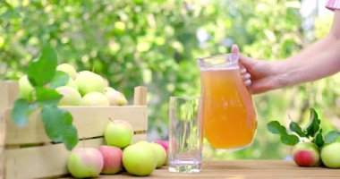 Kadınların eli, elma bahçesinde, bir sürahide bulunan elma suyunu bir bardağa döküyor. Olgun elmalar kutuda ve masada yatıyor.