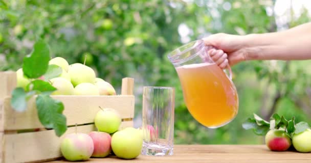 在夏天的苹果园里 女人的手正在把苹果汁从酒瓶里倒入木桌上的杯子里 成熟的苹果放在盒子和桌子上 — 图库视频影像