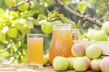 Bir kutu elma, cam ve bir sürahi taze elma suyu ahşap masa üzerinde bahçe arka planında. Elma hasatı. Meyve suyu üretimi.