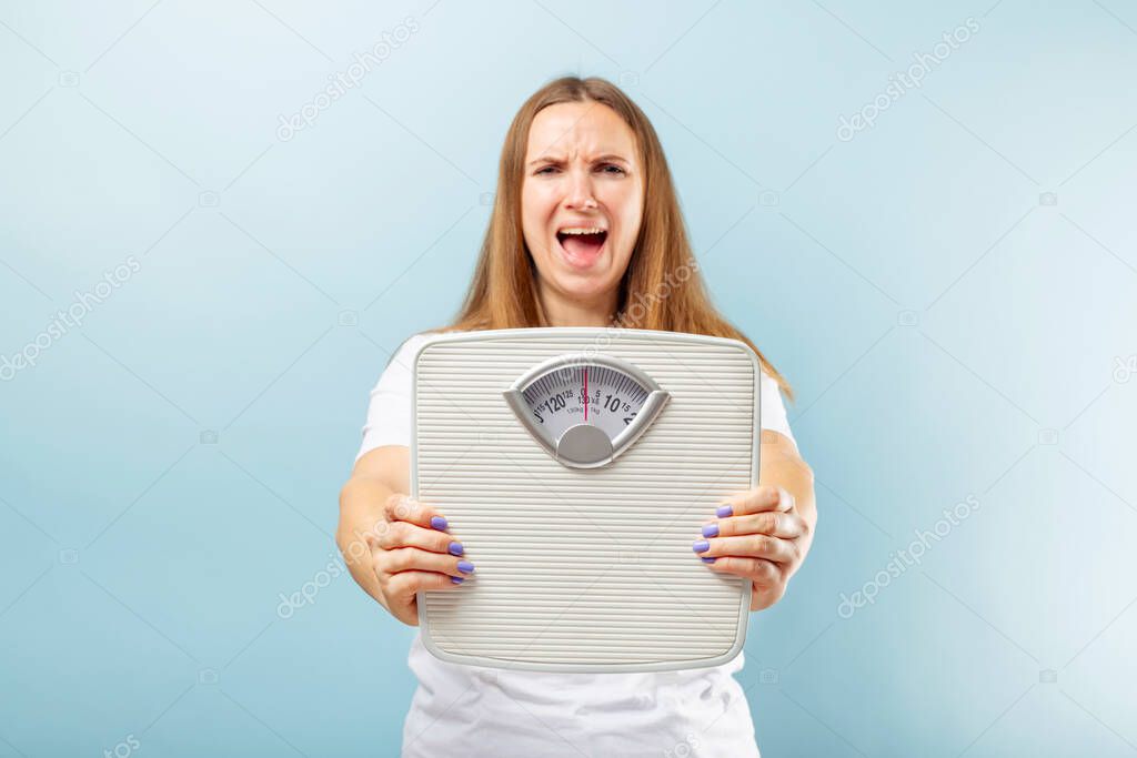 Frustrated young brunette woman in a white t-shirt with weight scale on blue background.